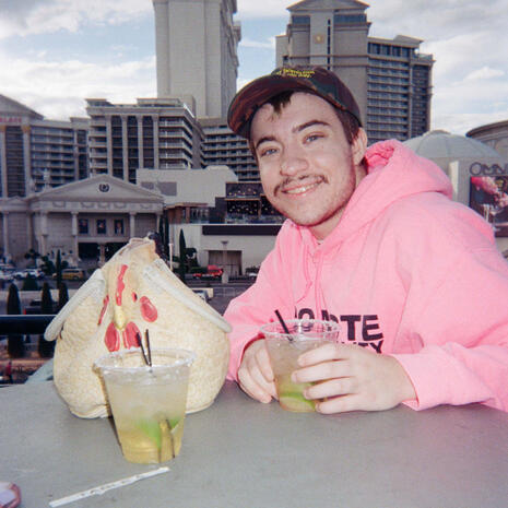 A transsexual in a pink hoodie & camo hat sitting with a rubber chicken purse, sharing a drink.